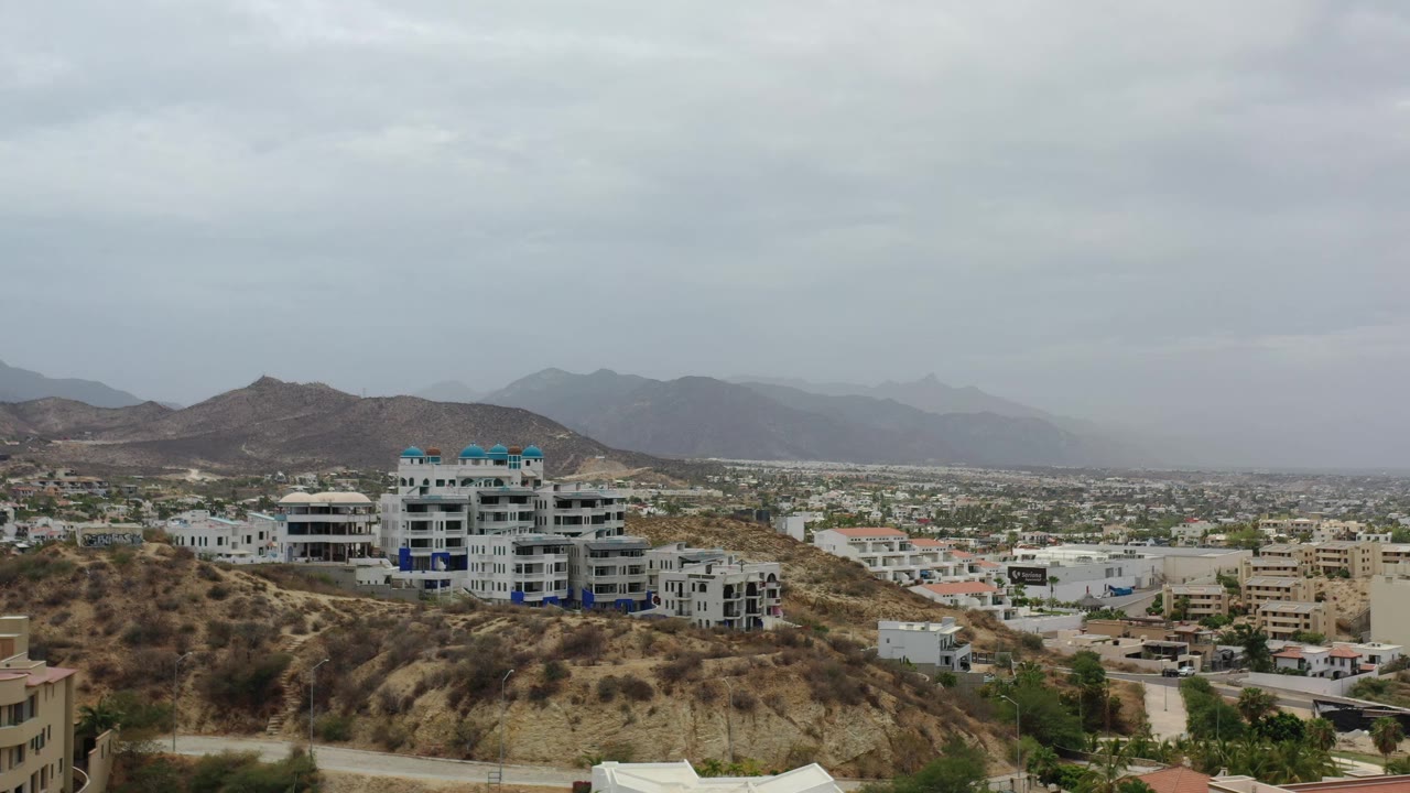 Wide Los Cabos hillside frame with a large white condo building near center, smaller houses below, a broad city plain, and muted mountains behind, no visible people.