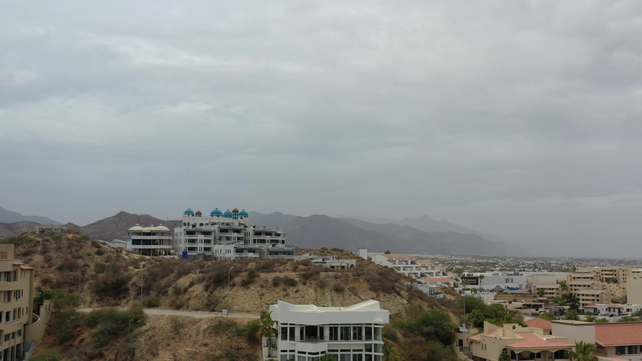 A wide Los Cabos hillside frame shows the ridge-top white buildings slightly more centered, with layered gray clouds, hazy mountains, and no visible people.