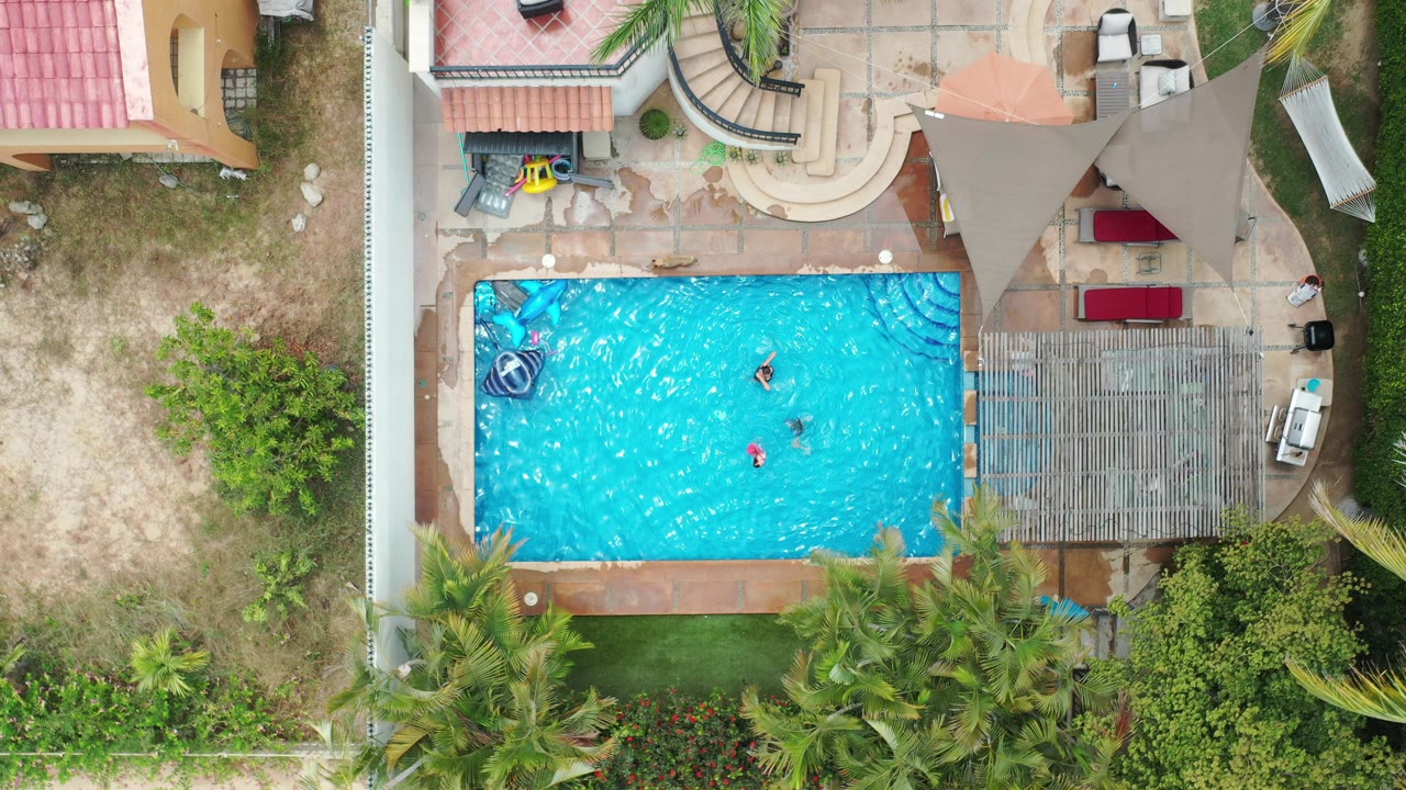 Overhead view of three people in a blue swimming pool beside patios and palms.