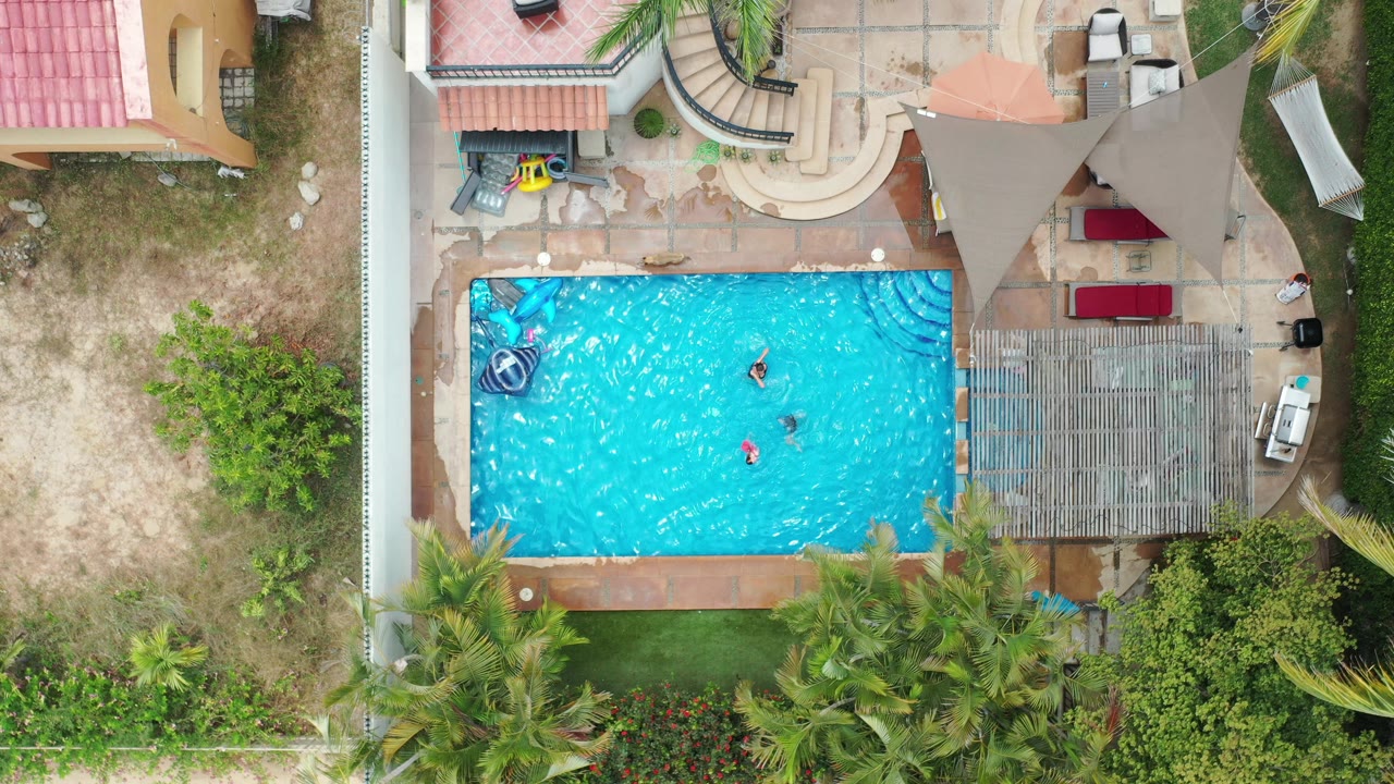 Overhead Los Cabos pool frame with the swimmers closer to the center of the water, a pool float near the left edge, and shaded patio furniture to the right.