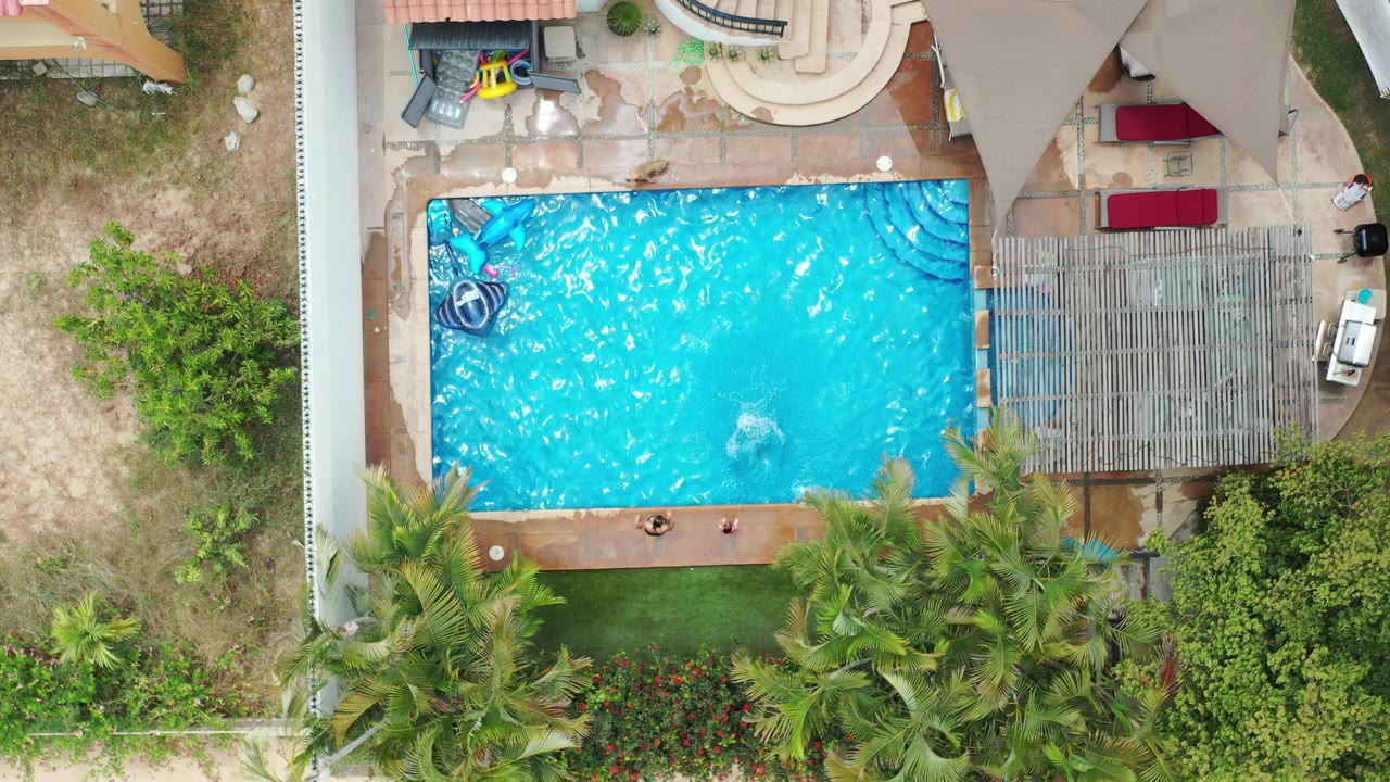 Overhead Los Cabos pool frame with sunlit ripples across the blue water, people along the lower pool edge, patio seating above, and dense green landscaping below.