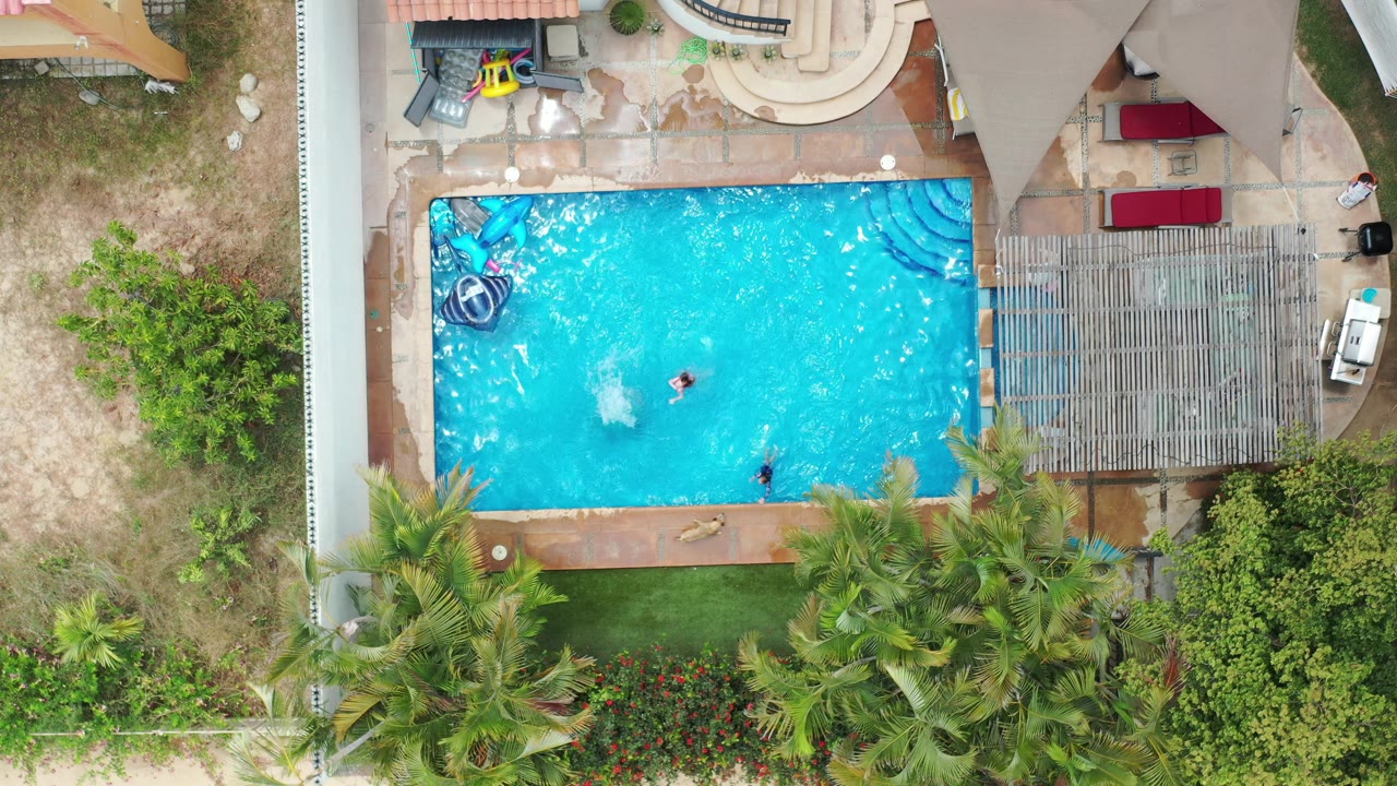 Top-down Los Cabos pool frame with two visible swimmers apart in the water, ripples across the bright blue surface, a shaded pergola on the right, lounge cushions above, and palms below.