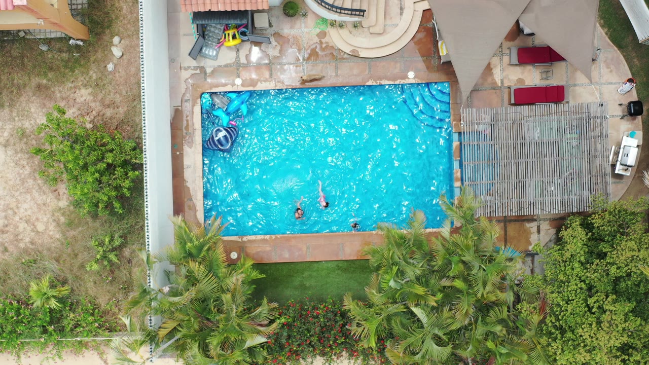 Aerial Los Cabos pool frame with children still visible near the center of the water, patio furniture and shade structure to the right, and garden greenery below.