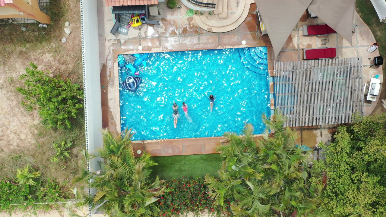 Overhead view in Los Cabos, Mexico, showing children spread across a bright blue backyard pool with floats, patio paving, palms, and a pergola around the water.