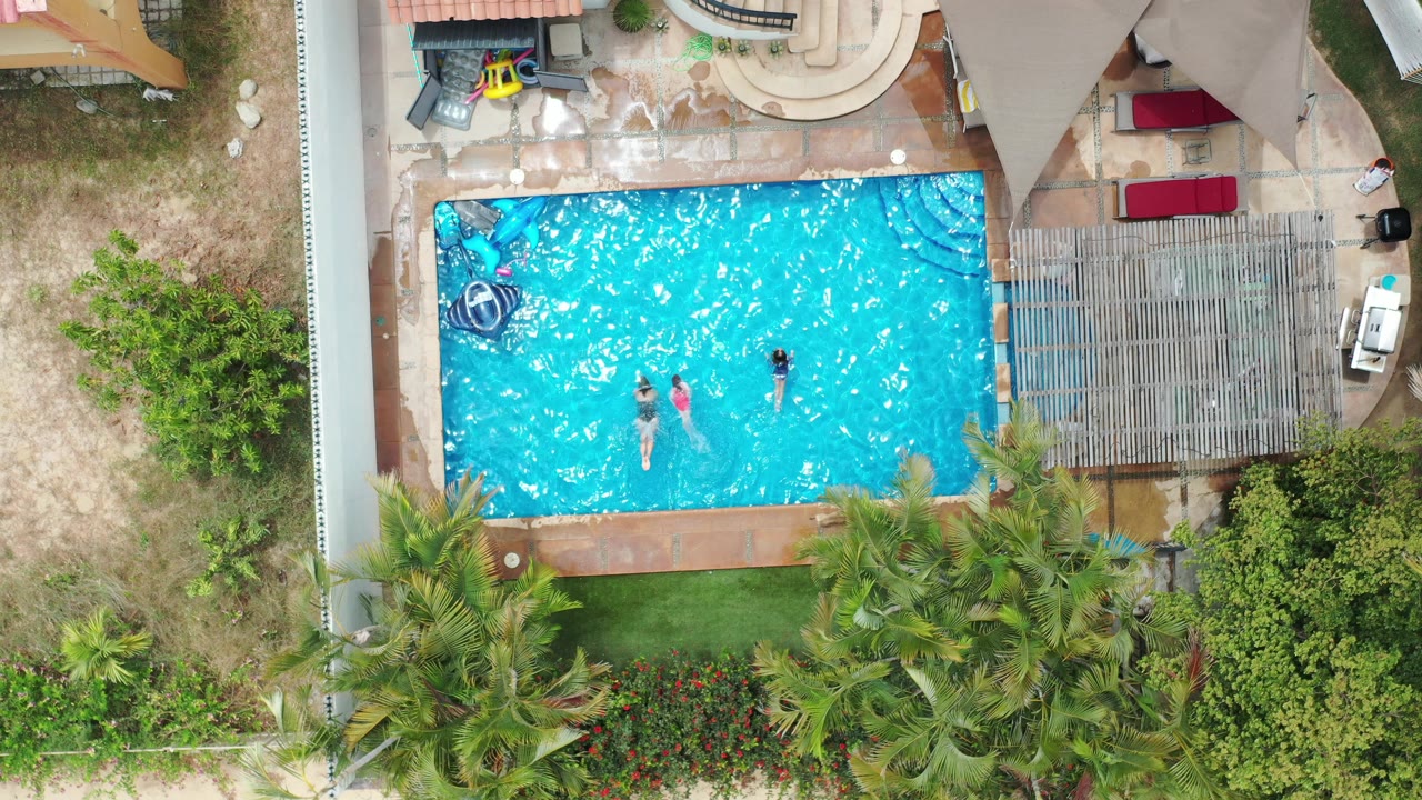 Overhead Los Cabos pool frame with children near the center of the water, a blue float at the upper left, red lounge cushions beside a pergola, and palms along the lower edge.