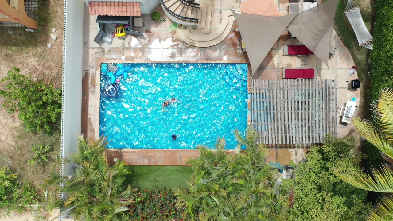 Overhead Los Cabos frame showing a bright rectangular pool, small swimmers, patio furniture, shade structures, and palms around a private home.