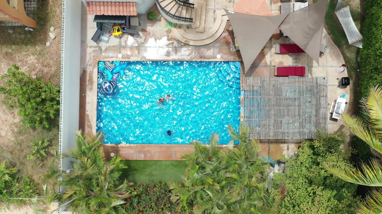 Straight-down Los Cabos pool view with two small people in the water, red lounge cushions, a pergola area, and dense greenery at the lower edge.