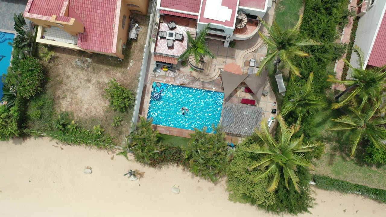 Aerial frame over Los Cabos, Mexico with the rectangular pool centered near beach sand, tiled roofs above it, dense palms to the right, and no visible people.