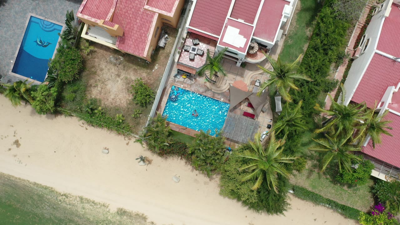 Overhead beach homes with red roofs, pools, palms, sand, and shallow water.