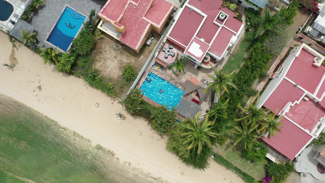 Overhead view of red-roofed houses, pools, sand, palms, and green coastal water.