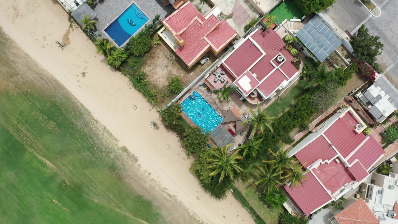 Overhead Los Cabos frame with green turf at left, a pale sandy strip, red-roofed homes, blue pools, palm trees, and no visible people.