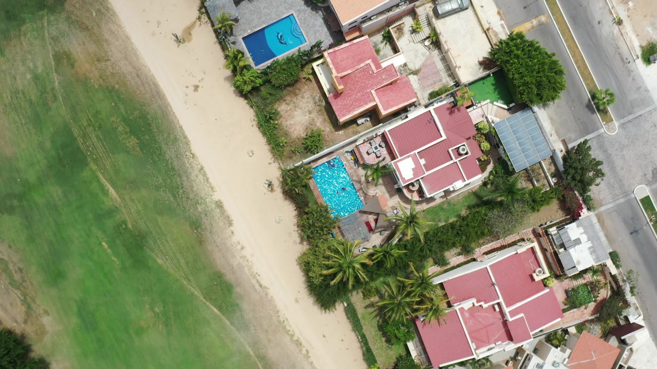 Overhead view of red-roofed homes, a blue pool, road, sand, and green ground.