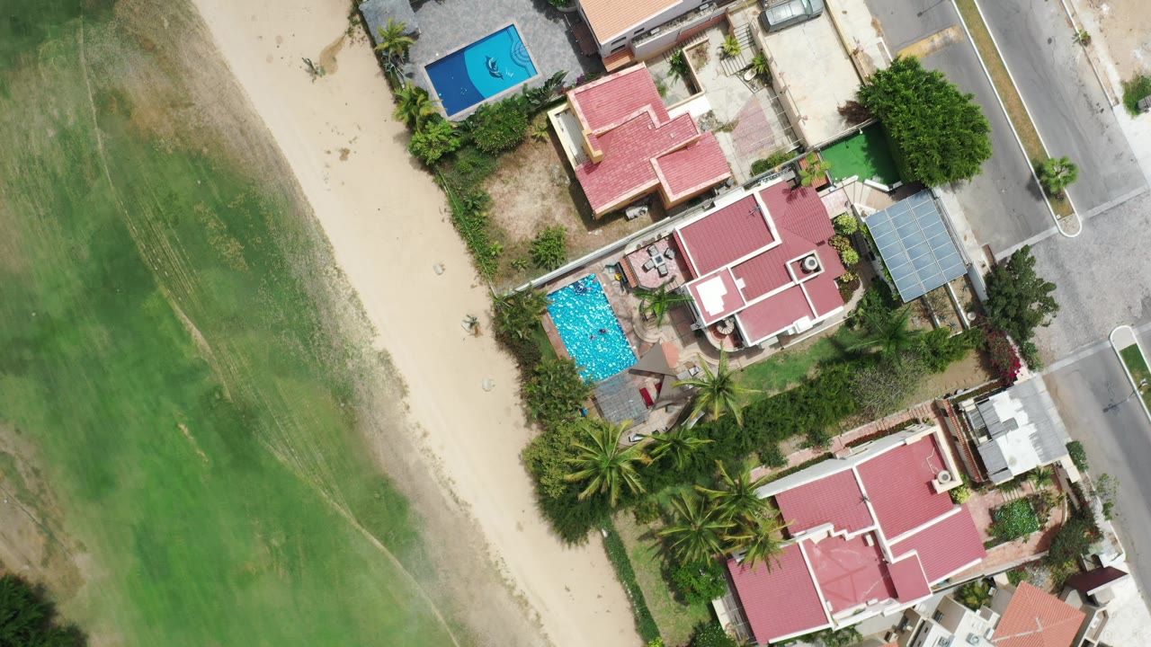 Top-down Los Cabos aerial frame with the blue pool and red-roofed homes shifted near center, bordered by a road on the right and green ground on the left, with no visible people.