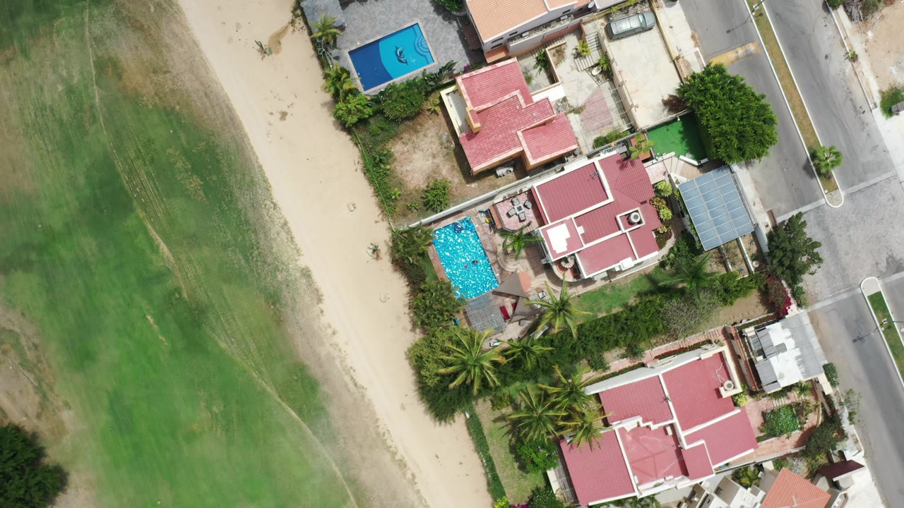 Aerial view of green open ground, sandy strip, houses, pool, and road.