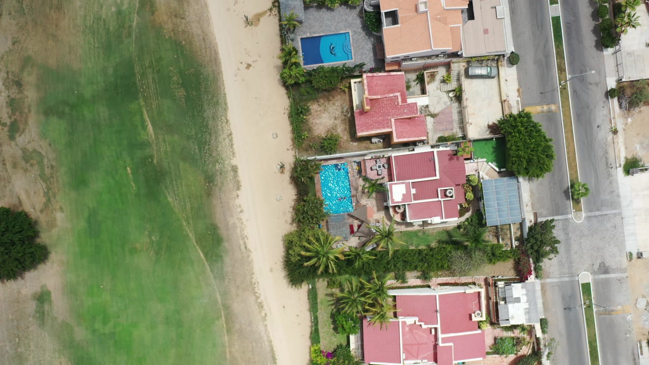 Straight-down aerial view in Los Cabos showing red-roofed homes, blue pools, palm trees, a paved road, sandy ground, green open land, and no visible people.