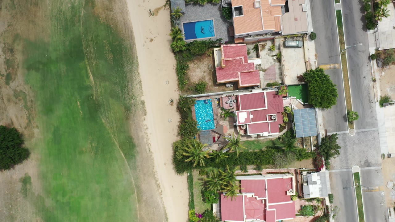 Overhead Los Cabos frame with tan and red roofs, two visible blue pools, palms, a vertical road edge, green land to the side, and no visible people.
