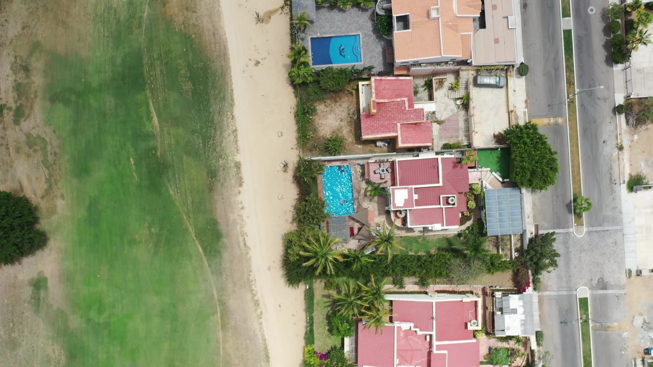 Top-down neighborhood view with pools, red roofs, palms, a street, and green open land.