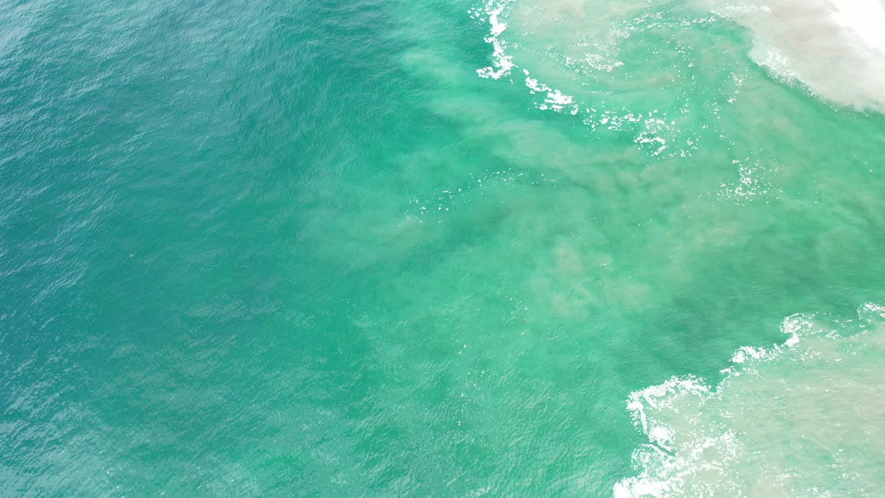 Overhead view of turquoise and blue ocean water in Los Cabos, Mexico, with white foam entering near the upper and lower right edges and no visible people.