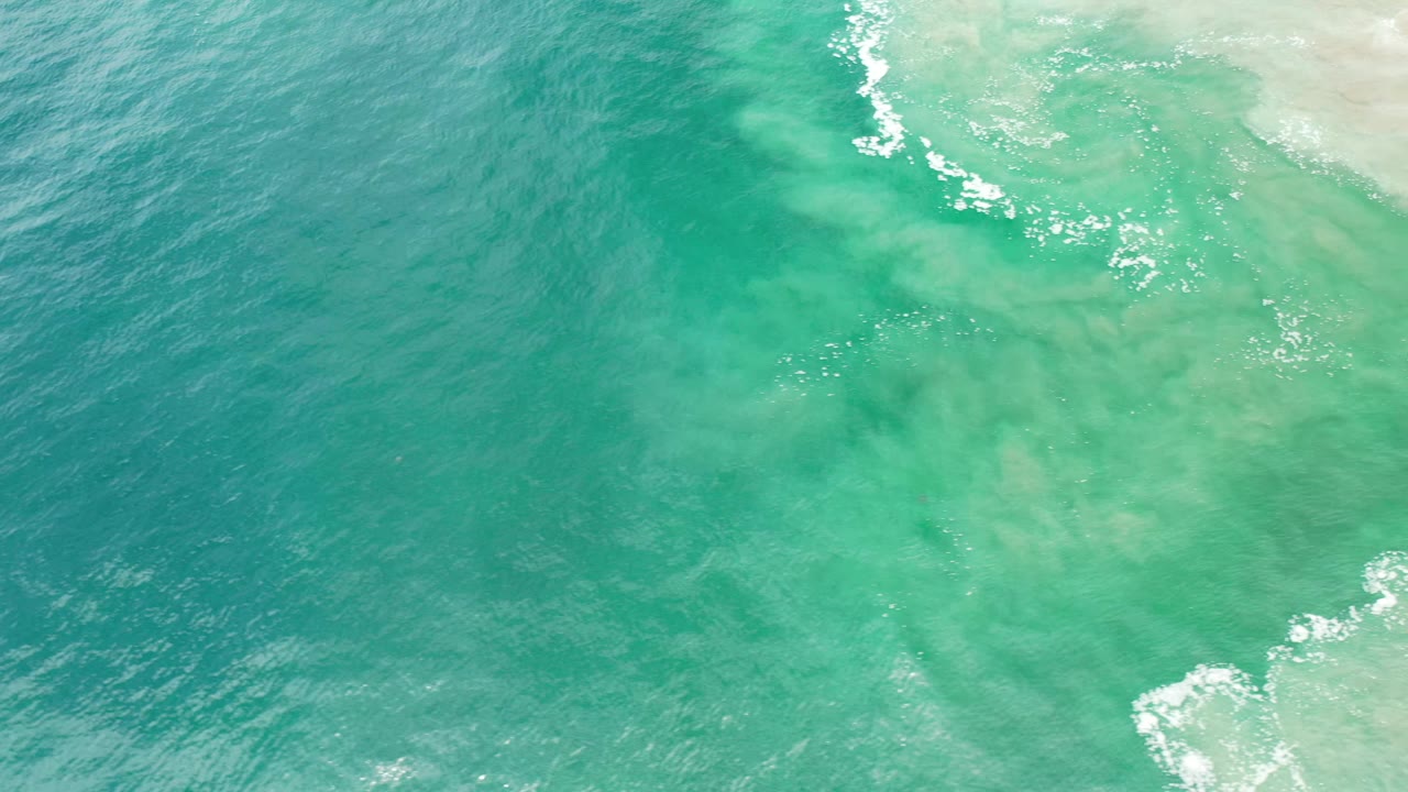 Los Cabos turquoise water shows ripples across the surface and a foamy wave edge entering from the upper right, with no visible people.