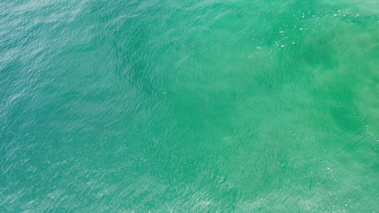 Overhead view of blue-green tropical water in Los Cabos, Mexico, with pale submerged patches, fine ripples, small sun glints, and no visible people.