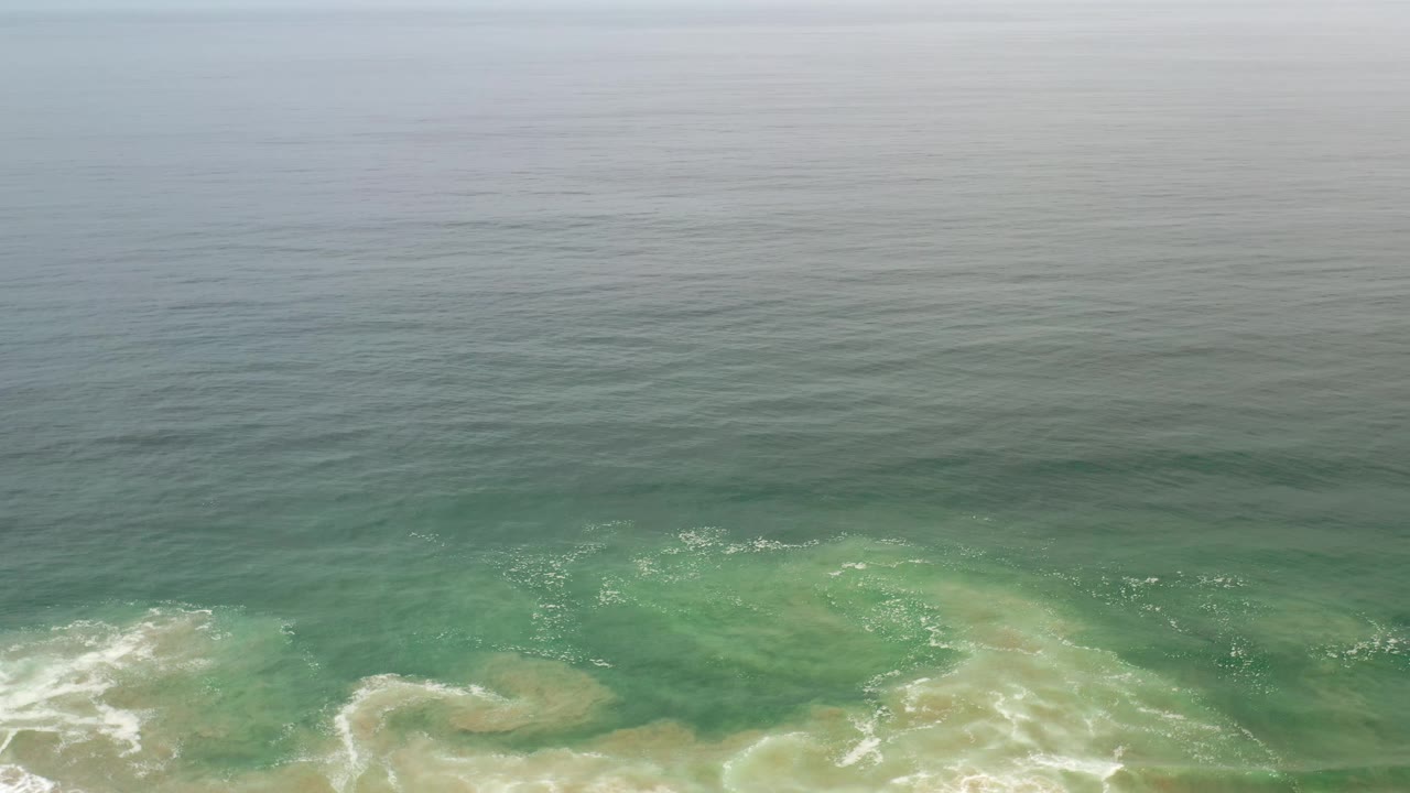 Aerial Los Cabos shoreline frame with darker offshore water above, green shallows below, pale sandy patches under the water, and no visible people.
