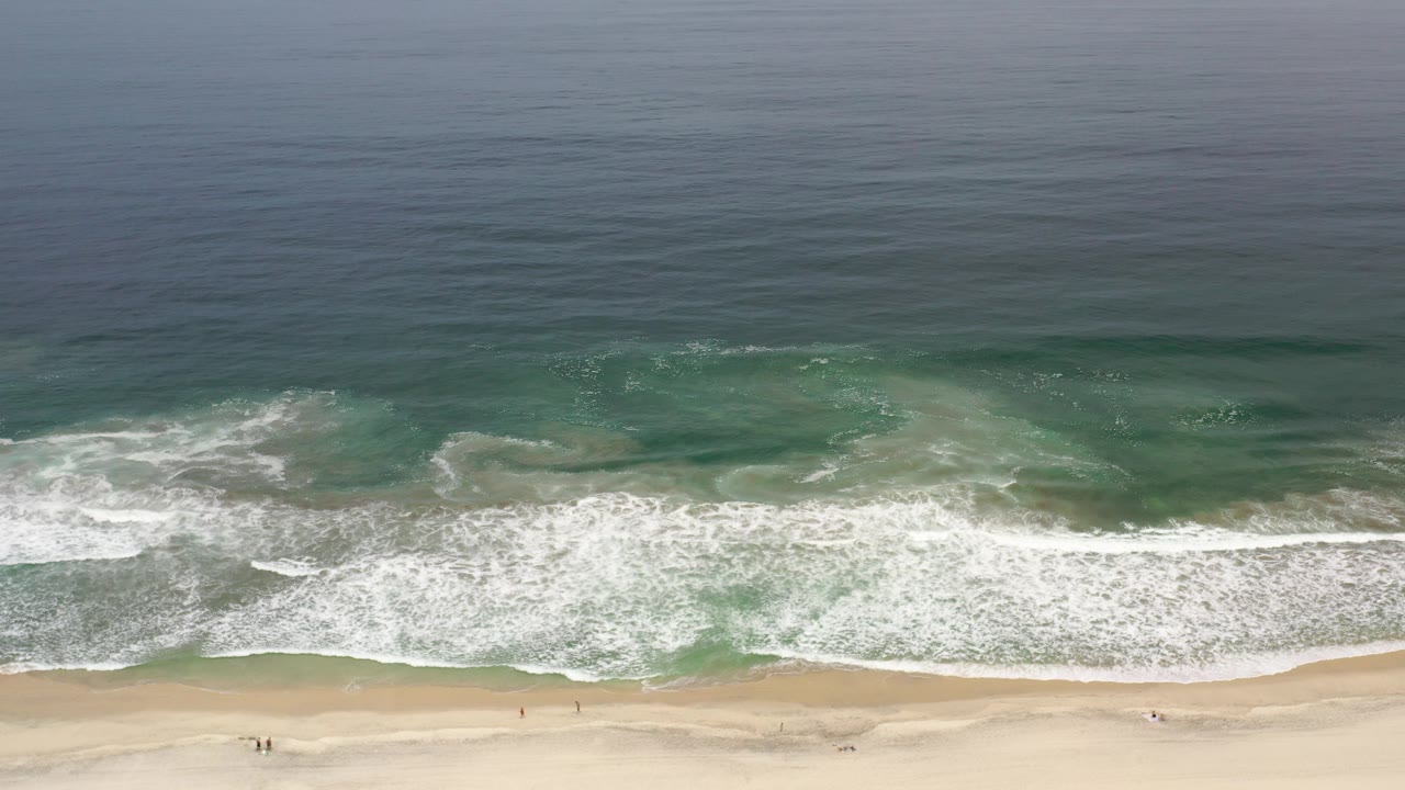 Opening aerial frame of a Los Cabos beach with pale sand below, white surf across the center, darker blue ocean above, and no visible people.