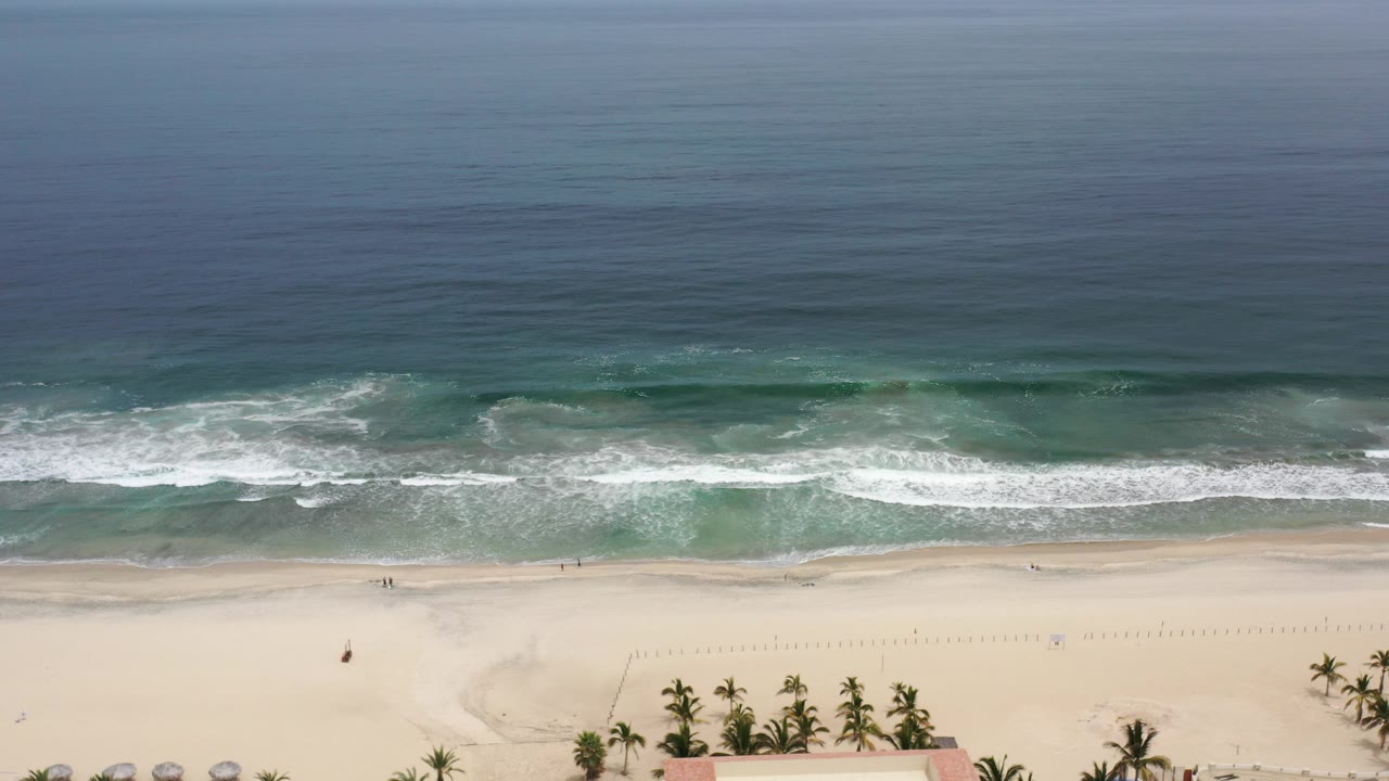 Aerial view of ocean surf reaching a wide sandy beach with palms at the bottom edge.