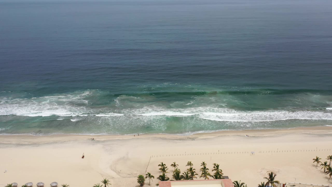 Los Cabos aerial beach frame with a broad band of whitewater crossing the sand line and palms clustered near the lower edge, no visible people.