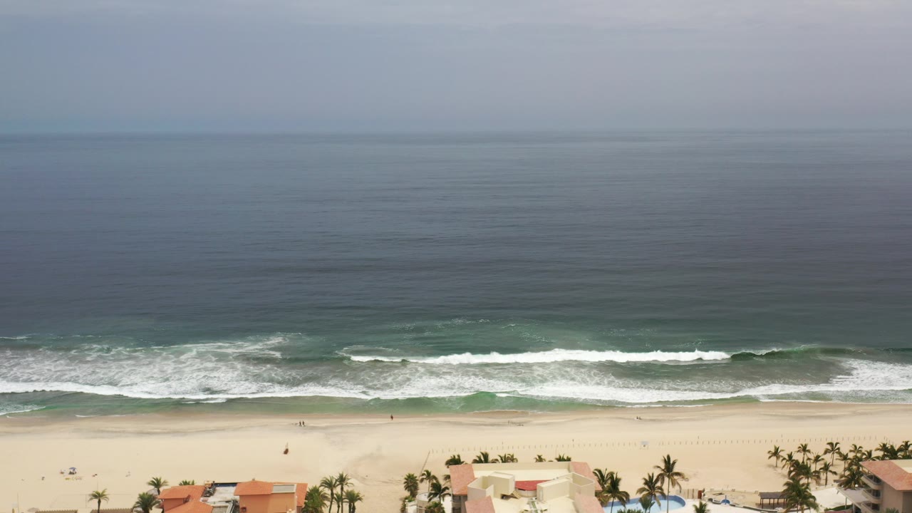 Elevated view of a Los Cabos beach with gray-blue ocean, white surf lines, pale sand, palms, and red-roofed coastal homes at the bottom edge, with no visible people.