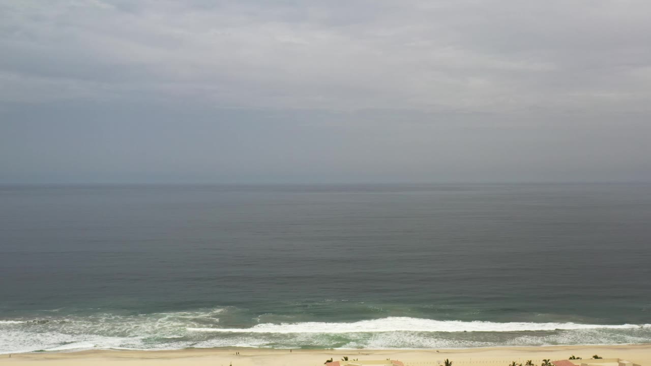Los Cabos shoreline seen from a high angle, with muted blue ocean, white wave bands, pale sand, and no visible people.