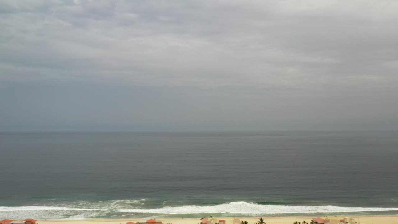 The Los Cabos oceanfront remains wide in frame, with a soft gray sky, horizontal surf bands, pale beach, and small rooftops along the lower edge with no visible people.