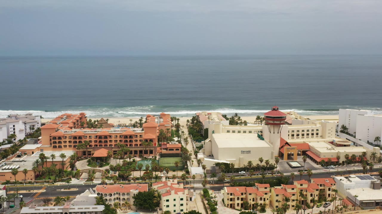 Los Cabos aerial view with a large orange-roofed building near center, white structures to the right, shoreline surf beyond, and no visible people.