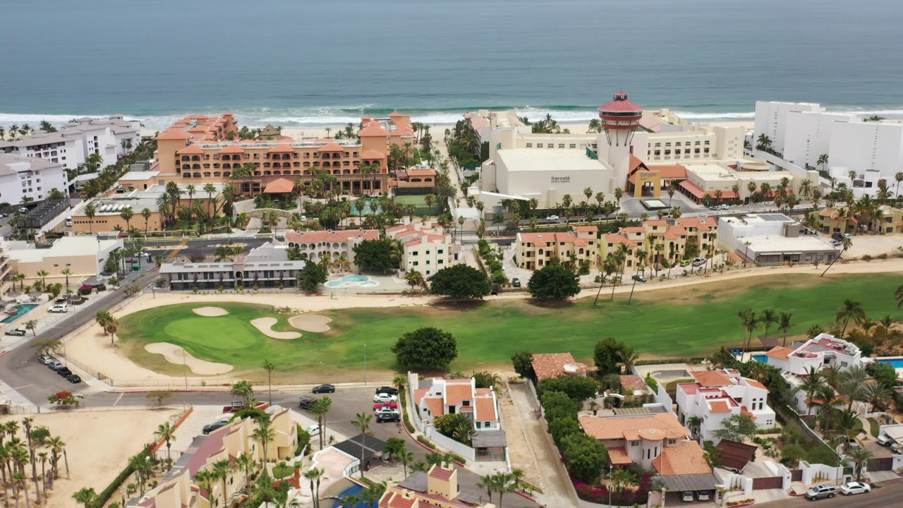 High aerial frame over Los Cabos with private homes in the foreground, a green golf course and resort buildings beyond, ocean surf in the background, and no visible people.