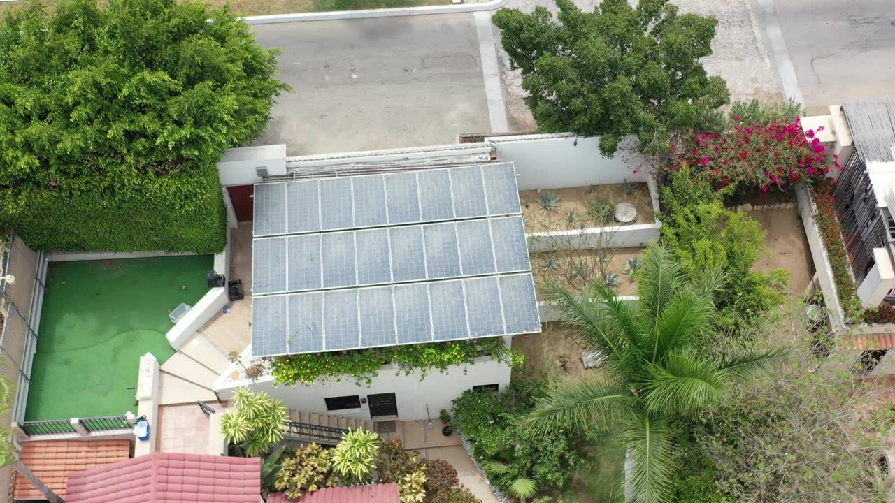 Overhead frame from Los Cabos, Mexico showing a white modern home with gridded roof panels, a green swimming pool at left, red tile roof in front, garden plantings, and no visible people.