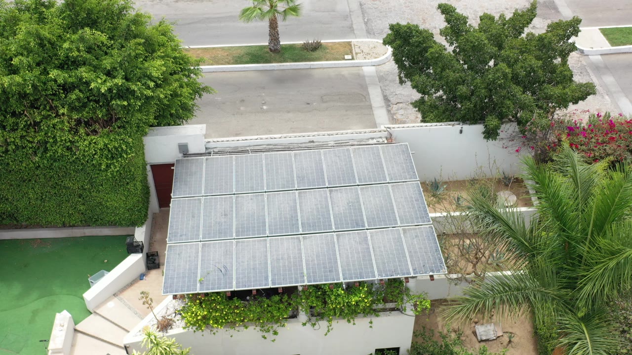 Opening frame in Los Cabos, Mexico showing a white residential exterior with a translucent canopy, hedges, palms, a green walkway area, and no visible people.