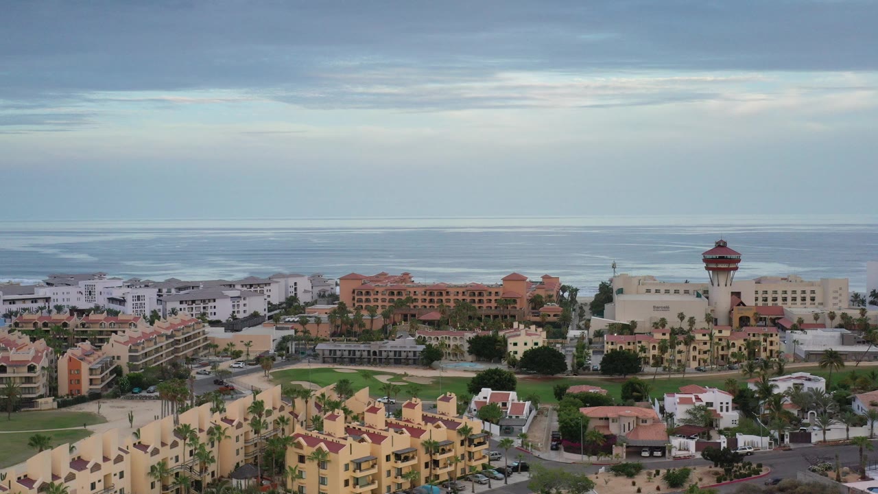 Wide elevated view in Los Cabos, Mexico, showing residences, green lawns, palms, and the ocean horizon under a cloudy sky, with no visible people.