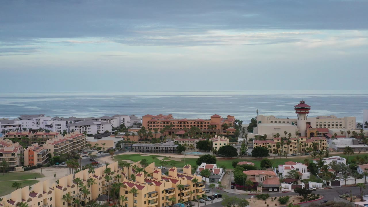 Elevated Los Cabos view with white and terracotta buildings, green landscaping, palms, and a muted ocean background, with no visible people.