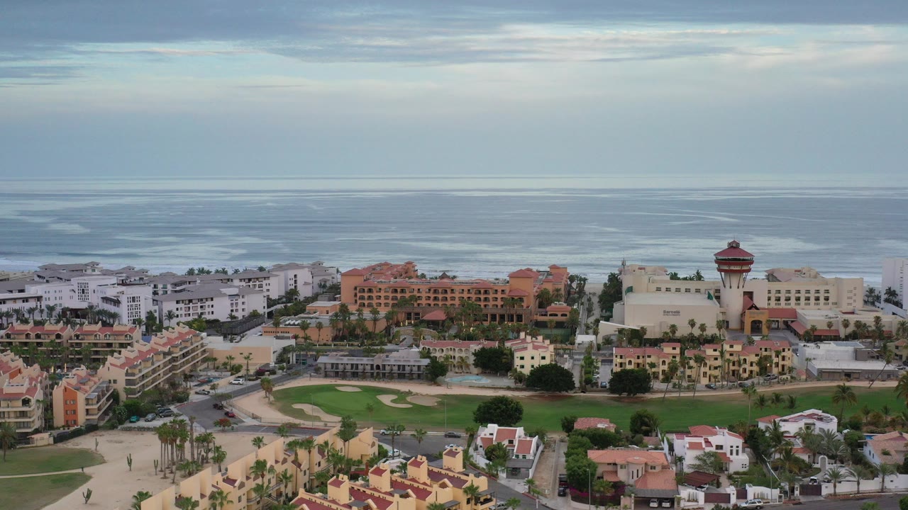 Wide aerial frame over Los Cabos, Mexico, showing seaside buildings, red roofs, a green landscaped area, ocean surf bands, cloudy sky, and no visible people.