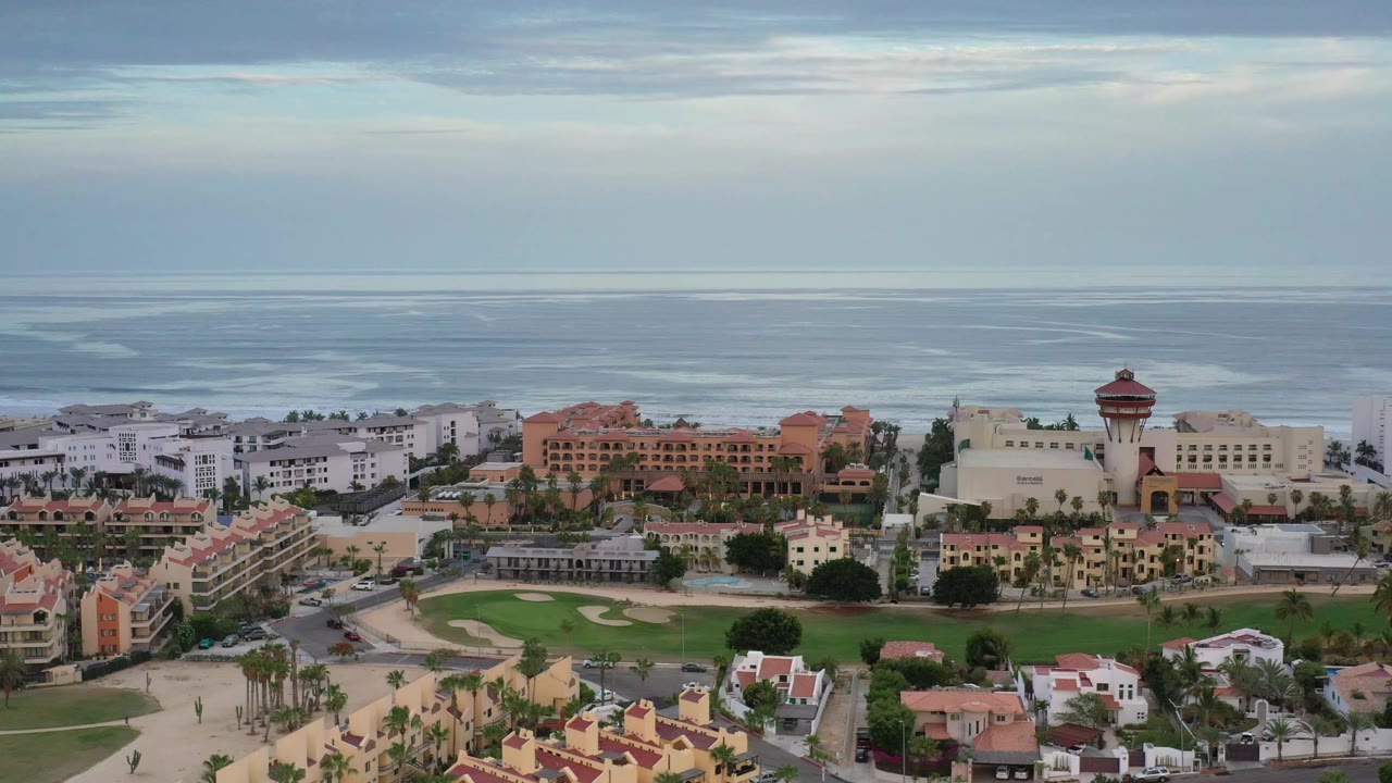 Aerial Los Cabos view with resort-style blocks across the lower frame, a green lawn area near center, the sea beyond, layered clouds above, and no visible people.