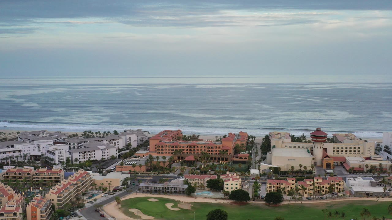 Aerial frame over Los Cabos, Mexico showing red-roof and white beachfront buildings below a wide blue-gray ocean with layered white surf and no visible people.