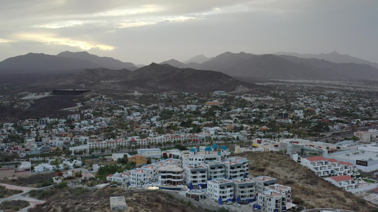 Opening aerial frame of Los Cabos, Mexico, with white buildings in the foreground, dark hills across the middle distance, hazy mountains behind them, and no visible people.