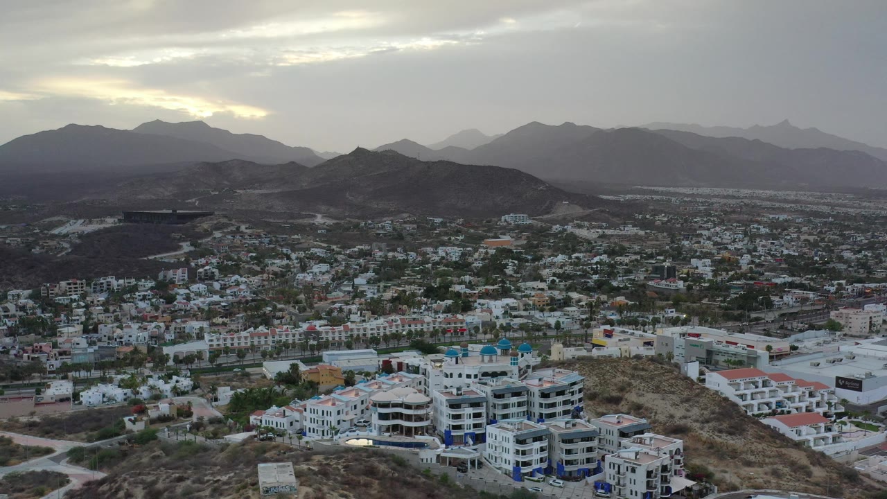 Early aerial view over Los Cabos, Mexico, showing a dense grid of pale buildings, a rounded hill near center, layered mountains in haze, and no visible people.