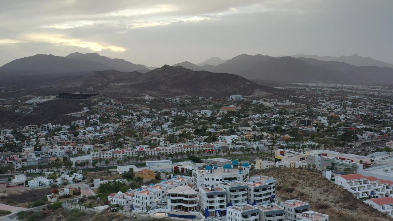 Los Cabos aerial view at 0:01 shows more of the town grid across the lower frame, a dark ridge in the center, hazy mountains beyond, and no visible people.