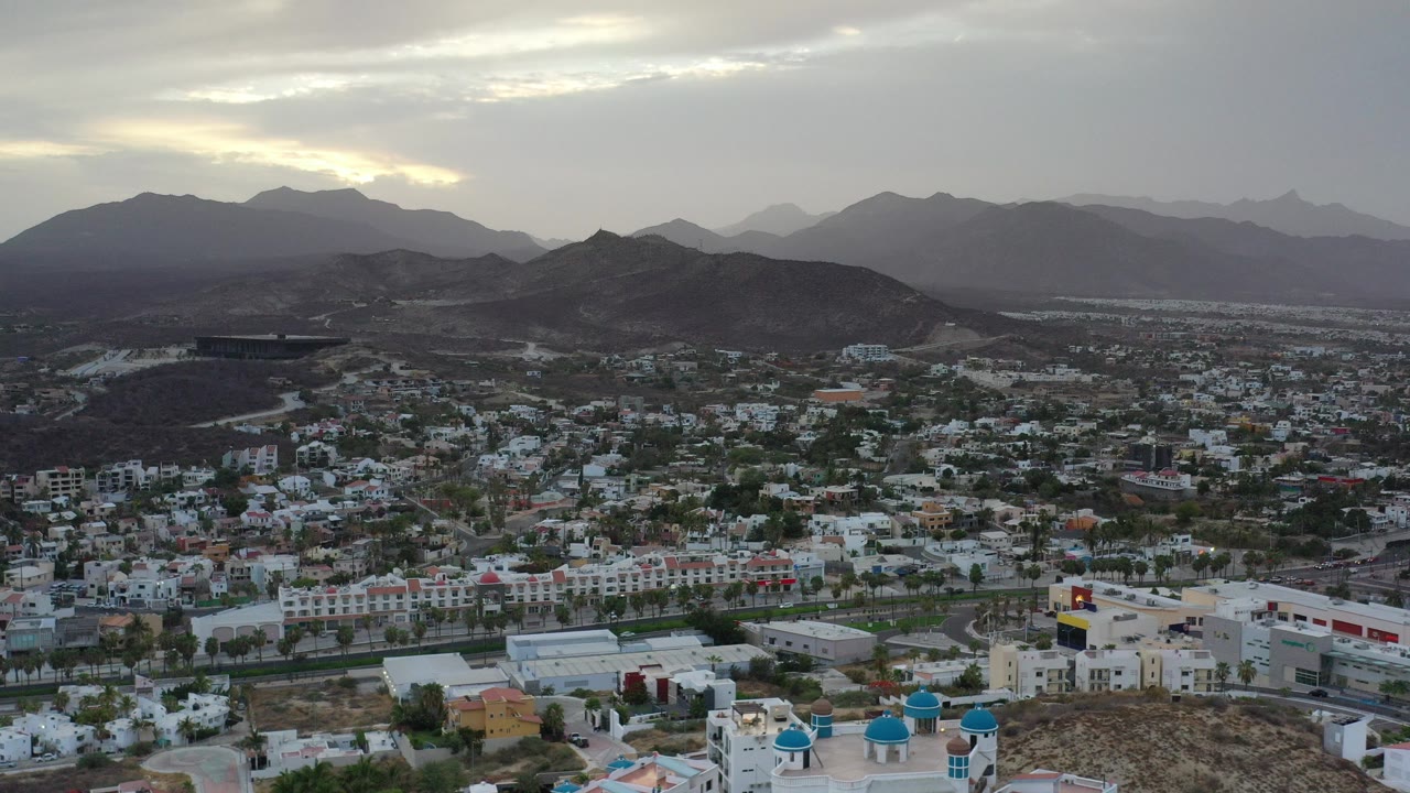 Wide aerial view of a city below dark hills and mountains under a cloudy sky.
