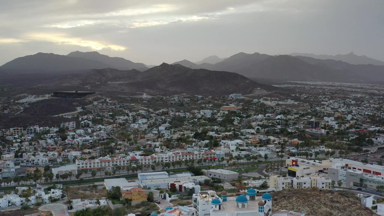 Los Cabos, Mexico appears from an elevated viewpoint, showing compact neighborhoods, a dark rounded hill, distant mountain ridges, muted gray light, and no visible people.