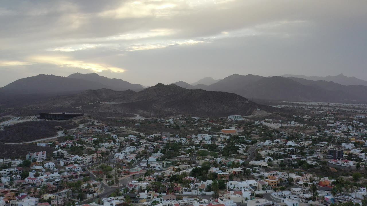 Aerial view of houses below dark hills and distant mountains under gray clouds.