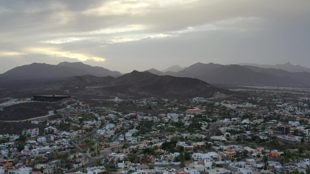 Los Cabos, Mexico view with dense low buildings in the foreground, a dark ridge through the middle, and hazy mountain layers beneath gray clouds, with no visible people.