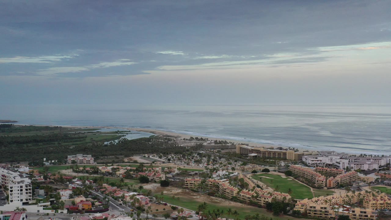 A high aerial view in Los Cabos, Mexico shows low-rise coastal buildings, roads, landscaped green space, beach surf, and a cloudy ocean horizon with no visible people.