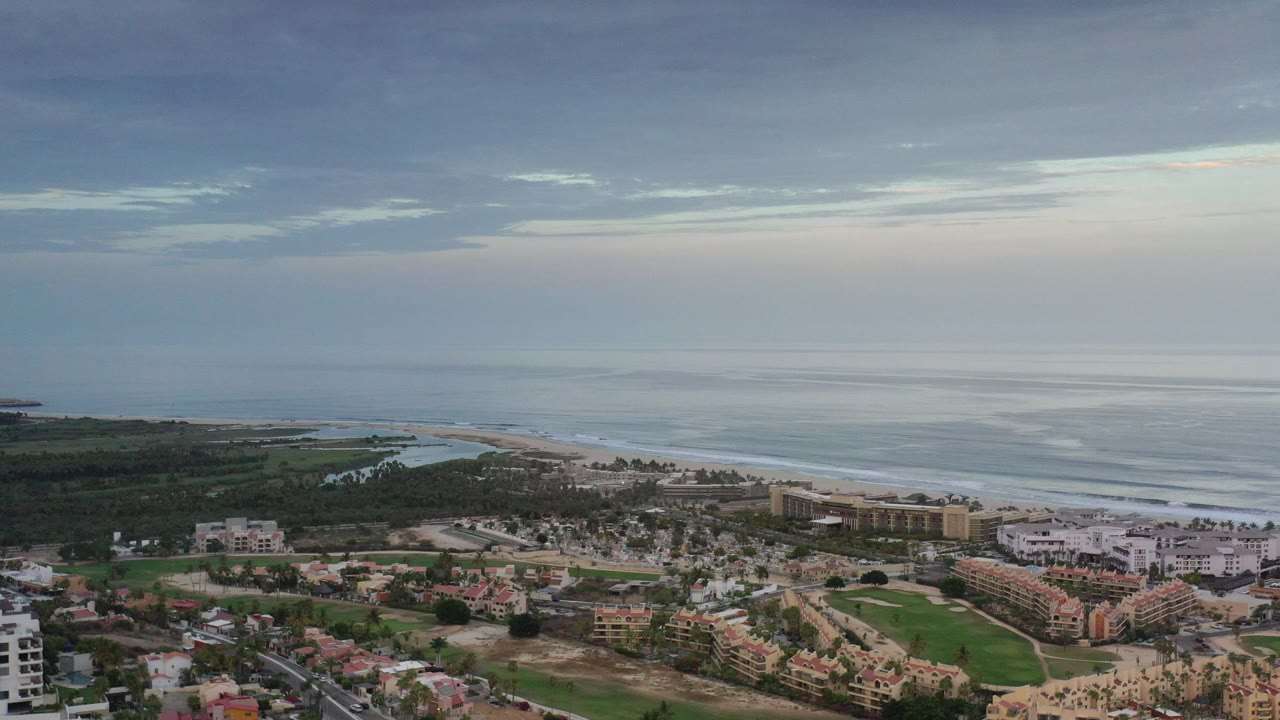 Opening aerial frame over Los Cabos, Mexico, with low-rise buildings below, surf beyond the beach, layered clouds above the horizon, and no visible people.