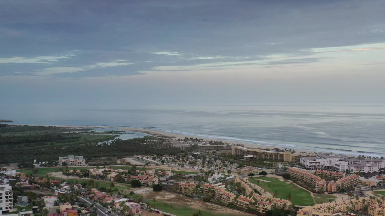 Aerial view of Los Cabos, Mexico, showing a green landscaped strip in the foreground, clustered buildings near the beach, smooth ocean water, and no visible people.